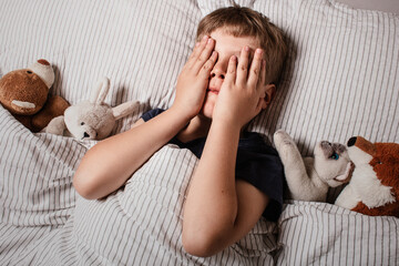 The boy was scared before going to bed. Night terrors in a child. The kid covers his face with his hands in a fear. Children's experiences. Boy in the bed. View from above. Sad psychological state.