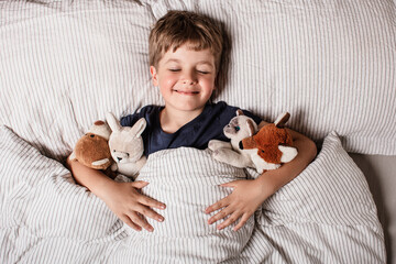 Happy child waking up in bed with big smile and raised arm, surrounded by stuffed animals. Joyful...