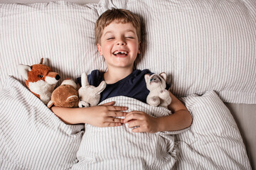 Happy child waking up in bed with big smile and raised arm, surrounded by stuffed animals. Joyful morning energy, childhood happiness and cozy home atmosphere. Fun before a sleeping. Kid smile in bed.
