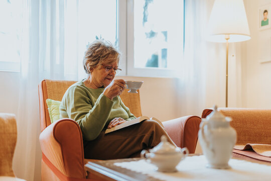 A serene scene of a senior woman sitting with a book and tea in a brightly lit, comfortable living room.