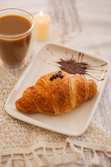 Breakfast scene with croissant and coffee. Breakfast setting featuring a golden croissant on a decorative plate with coffee and a soft candlelight glow. Selective focus.