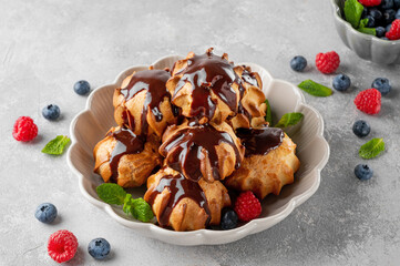 Profiteroles with vanilla cream, fresh berries and chocolate glazed in a bowl on a gray concrete background. A delicious French dessert.