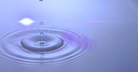 Hovering water droplet creating concentric ripples on calm water surface in studio, with lens flare