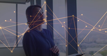 Standing woman wearing blue blazer near office window, holding tablet with digital network overlay