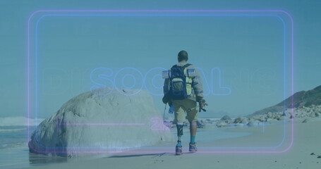 Walking man with prosthetic leg along sandy shoreline, with backpack, board and camera tripod