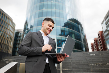Business professional engages with laptop, smiling in modern outdoor urban setting amidst sleek glass buildings