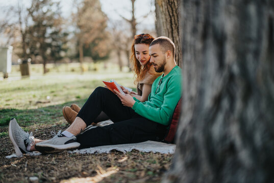 A couple is relaxing together outdoors, sitting on a blanket under a tree, sharing a book, emphasizing leisure, connection, and the peaceful outdoors.