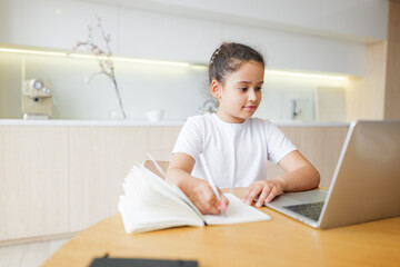 Young girl engaged in online learning, balancing note-taking and digital education in a modern kitchen setting, fostering creativity and growth