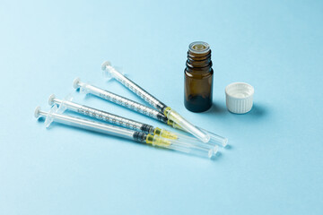 Opened brown medicine vial with white cap next to several disposable syringes on a light blue...