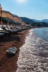city beach with sun loungers and umbrellas with beautiful coastline and view of Adriatic sea in Petrovac, Montenegro, with high forested hills in the background