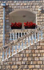 a beautiful balcony in the form of an arch and a steps with balustrades is decorated with flowerpots, a stone decorative wall is illuminated by bright sunlight