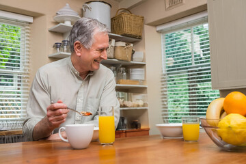 Senior man scooping breakfast from bowl with spoon at kitchen counter with mug, juice and fruit