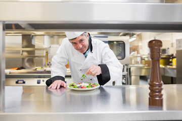 Male chef in uniform garnishing salad with herb sprig on stainless counter with pepper mill, ovens