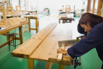 Asian female woodworker guiding plank through table saw in navy work jacket at carpentry workshop