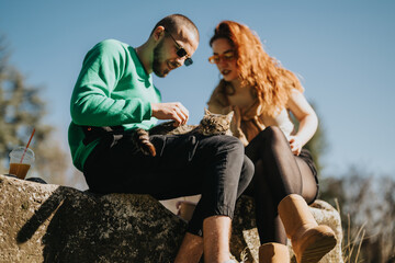A young couple enjoying their leisure time sitting on a rock with a cat, basking in the outdoor...