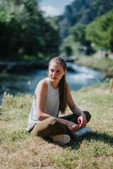 A serene moment depicting a young woman seated on the grass next to a calm river surrounded by lush...