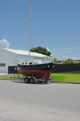 Sailboat on Trailer Parked Outdoors on a Sunny Day Near a Warehouse. Red and black hull on a trailer parked outdoors on a sunny day, surrounded by greenery and buildings, depicting outdoor and maritim