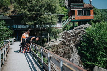 A group of friends standing on a bridge, enjoying and observing the natural surroundings by a scenic area featuring rustic cabins and greenery, under bright summer sunlight.