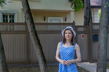 Elegant young woman in polka dot dress and white hat posing outdoors near residential fence