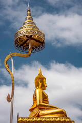 A golden Buddha statue in a serene, meditative pose, against a backdrop of a blue sky with white...