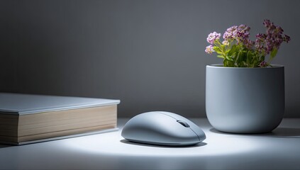 A gray computer mouse sits on a white surface, illuminated by a soft light, beside a book and a small potted plant
