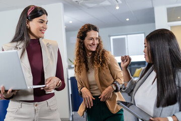 Diverse women coworkers wearing blazers collaborating at workstations using laptop and tablet