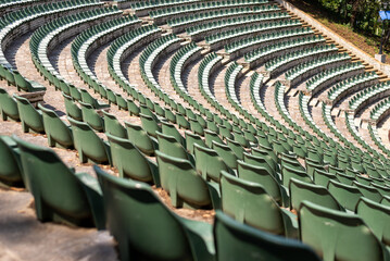 Large, open-air stone amphitheater is filled with numerous rows of green plastic seats arranged in a semi-circular pattern