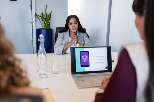 Diverse female colleagues in business attire talking app mockup in meeting room with laptop and pen