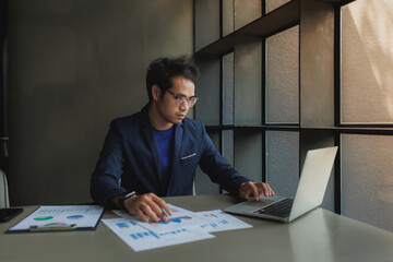 An Asian businessman sits tensely at his desk in a modern office, surrounded by documents and a...
