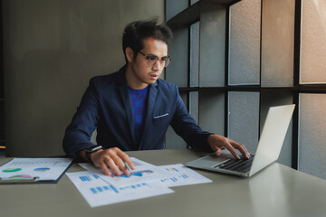 An Asian businessman sits tensely at his desk in a modern office, surrounded by documents and a...