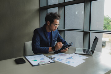 An Asian businessman sits tensely at his desk in a modern office, surrounded by documents and a...