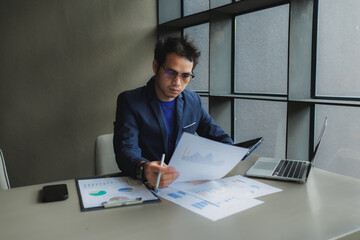 An Asian businessman sits tensely at his desk in a modern office, surrounded by documents and a...
