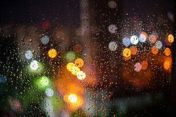 A window covered in raindrops and blurred view of colorful bokeh night city lights outside in rainy evening. Abstract background