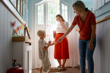 Mother saying goodbye to her son at preschool entrance as the teacher warmly guides him into the...