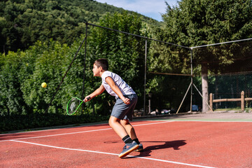 Fototapeta premium Boy playing tennis on a vibrant red court, preparing to strike a ball with his racket, immersed in the beauty of nature