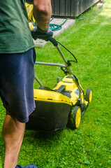 Rear view of man in shorts cutting grass with electric mower in backyard. Summer gardening and lawn care concept. High quality photo