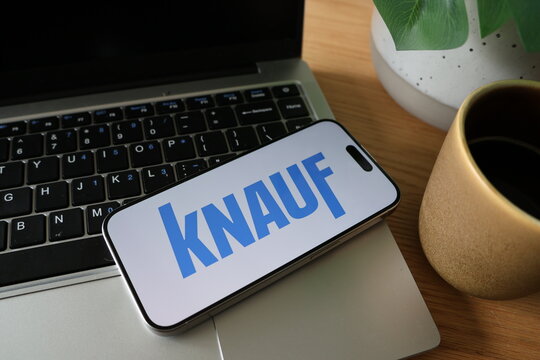 Kiel, SH, Germany, 08-07-2025: Close-up of a smartphone displaying the KNAUF logo on a laptop keyboard, accompanied by a potted plant and a coffee cup