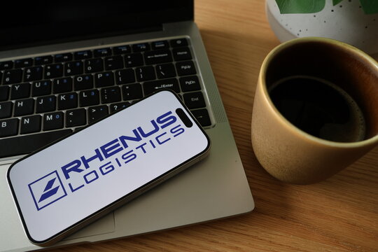Kiel, SH, Germany, 08-07-2025: A smartphone displaying the Rhenus Logistics logo rests next to a laptop and a cup of coffee on a wooden table