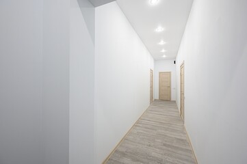 well-lit hallway with white walls and a light gray wood-patterned floor. There are three wooden doors evenly spaced along the hallway. Recessed lighting illuminates the space