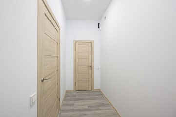 well-lit hallway with white walls and a light gray wood-patterned floor. There are three wooden doors evenly spaced along the hallway. Recessed lighting illuminates the space