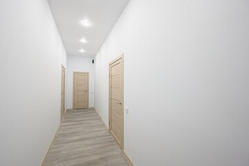 well-lit hallway with white walls and a light gray wood-patterned floor. There are three wooden doors evenly spaced along the hallway. Recessed lighting illuminates the space