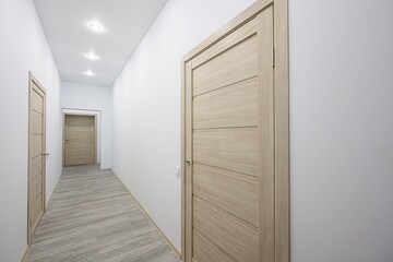 well-lit hallway with white walls and a light gray wood-patterned floor. There are three wooden doors evenly spaced along the hallway. Recessed lighting illuminates the space