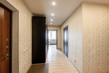 well-lit hallway with patterned wallpaper. A dark wardrobe stands on the left, and a doorway leads to another room in the distance. A wooden door is on the left