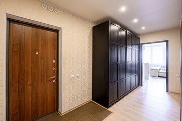 well-lit hallway features a wooden door, a dark wardrobe, textured wallpaper, and a glimpse into a bright room through an open doorway
