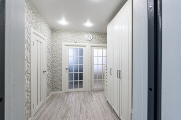 bright hallway with white doors, patterned wallpaper, wood-like flooring, a white clock, and a white wardrobe