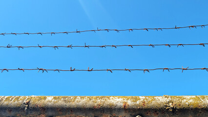 Barbed wire fence stretches across a clear blue sky, creating a stark contrast with the weathered...