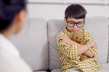 Asian boy with ADHD sitting alone on sofa hugging teddy bear displaying sadness and emotional...