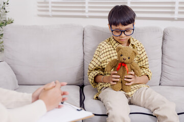 Asian boy seated on sofa holding teddy bear with shy smile during therapy session while female...