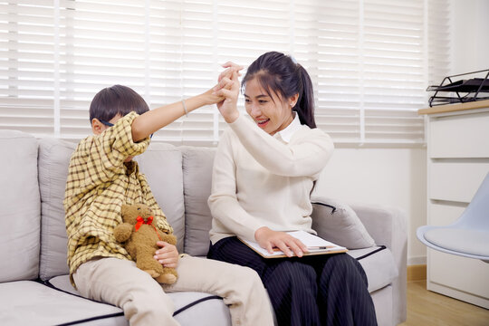 Asian boy holding teddy bear while interacting with female psychologist during playful therapy session, engaging in high-five gesture to strengthen bond and improve emotional expression - Powered by Adobe