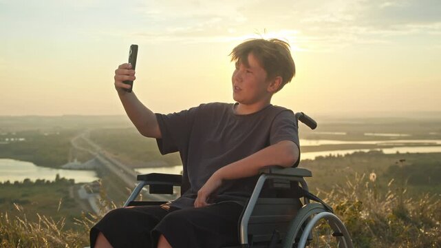 A boy in a wheelchair, against the backdrop of a summer sunrise, is talking on a video call using his phone. He waves his hand amicably, and his interlocutor greets him.
